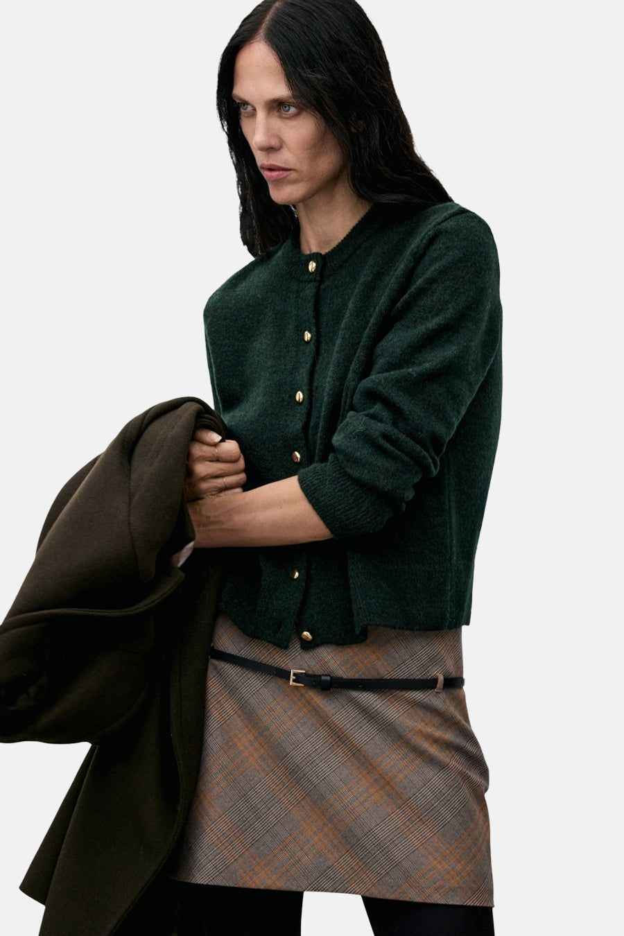 Cardigan vert à boutons dorés de MANGO, porté avec une jupe à carreaux marron et une ceinture fine noire.
