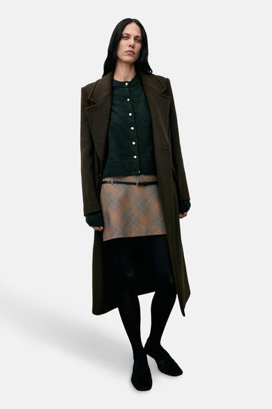 Cardigan vert MANGO porté avec une jupe à carreaux, un manteau long et des collants noirs.