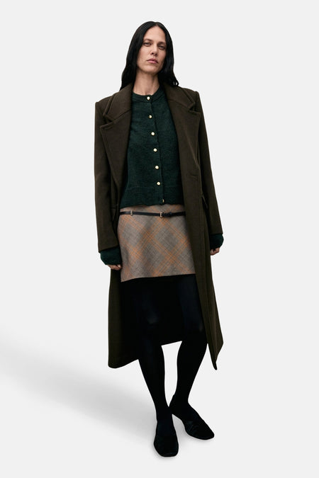 Cardigan vert MANGO porté avec une jupe à carreaux, un manteau long et des collants noirs.