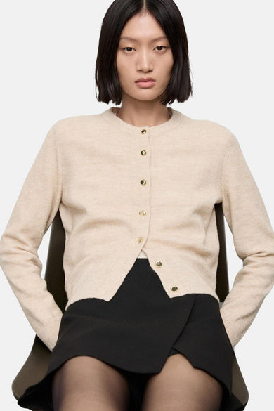 Cardigan beige en maille de MANGO, avec boutons dorés, porté sur une jupe noire et des collants noirs.
