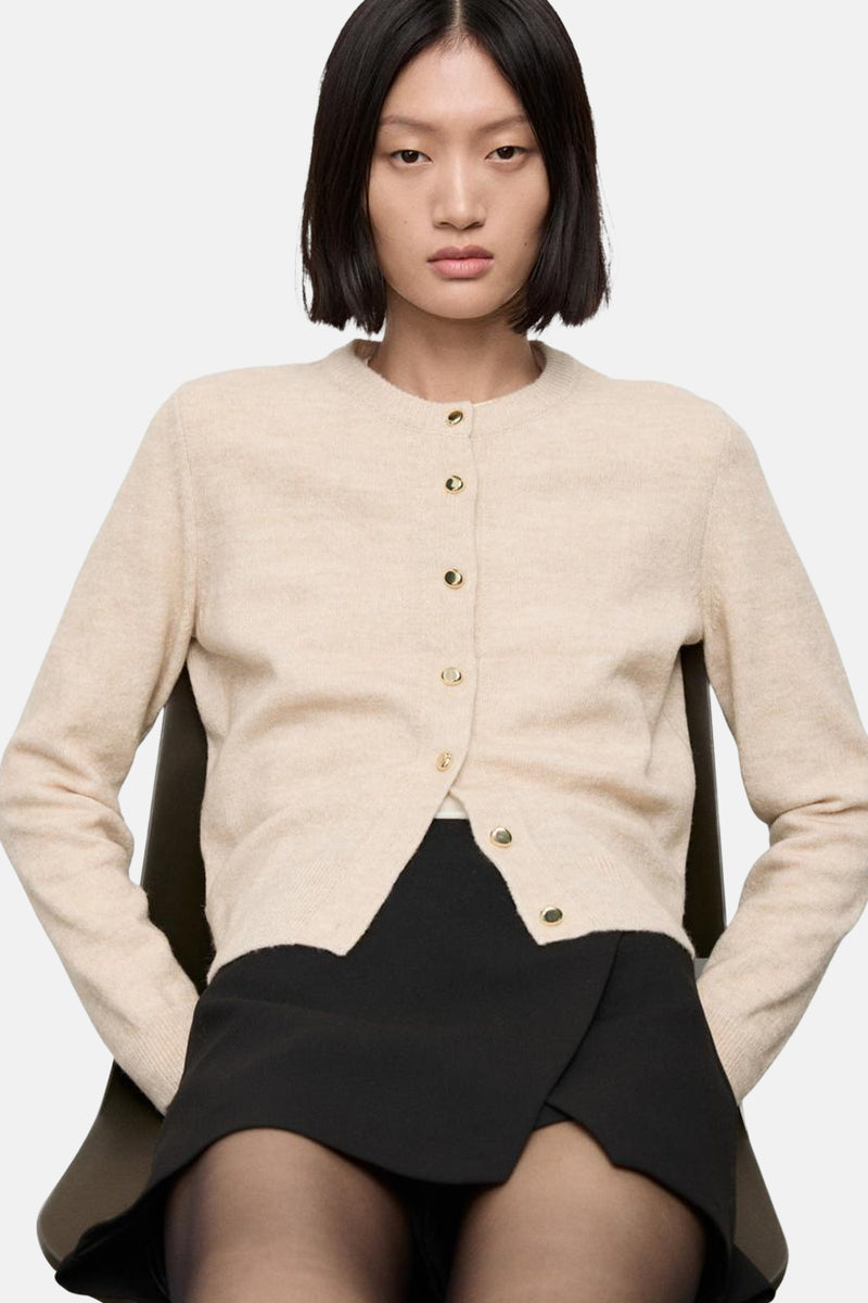 Cardigan beige en maille de MANGO, avec boutons dorés, porté sur une jupe noire et des collants noirs.
