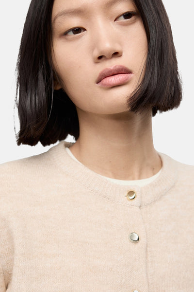 Cardigan beige en maille de MANGO, avec des boutons dorés, porté avec un t-shirt blanc.
