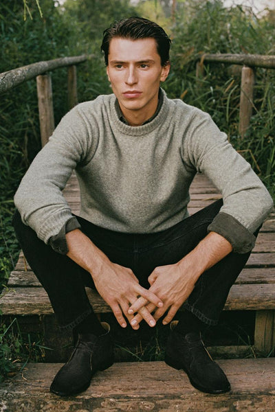 Pull col rond vert de MANGO, porté avec un pantalon noir et des bottines en daim, assis sur un pont en bois.
