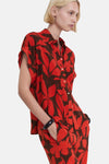 Chemisier rouge à manches courtes de MANGO, avec un imprimé floral marron et porté avec un bracelet noir.
