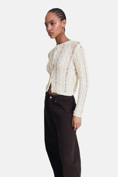 Ecru gebreid vest met open structuur van MANGO, gedragen met een bruine broek met hoge taille.