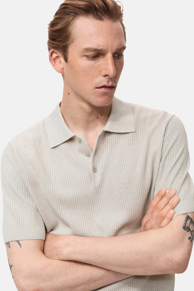 Polo écru à manches courtes de MANGO, en maille côtelée texturée, porté par un homme aux bras croisés.
