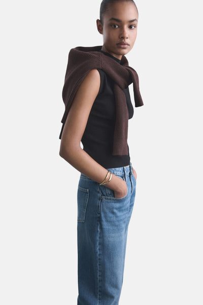 Jean en denim bleu moyen de MANGO, porté avec un pull marron sur les épaules et un bracelet doré au poignet.