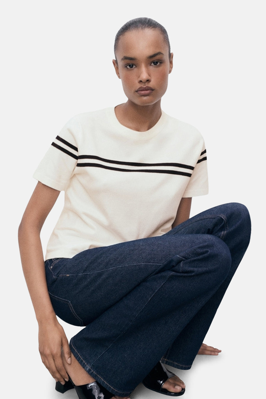 T-shirt blanc à manches courtes de MANGO, avec deux rayures horizontales noires sur le buste, porté avec un jean bleu.
