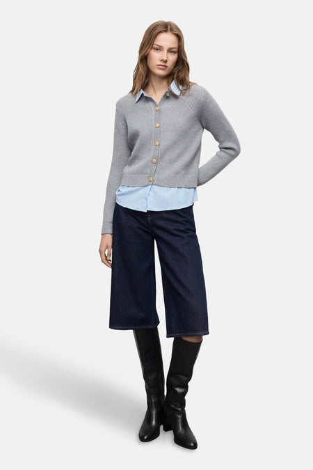 Grijze cardigan van MANGO, gedragen over een lichtblauw gestreept overhemd, met een wijde spijkerbroek en zwarte laarzen.