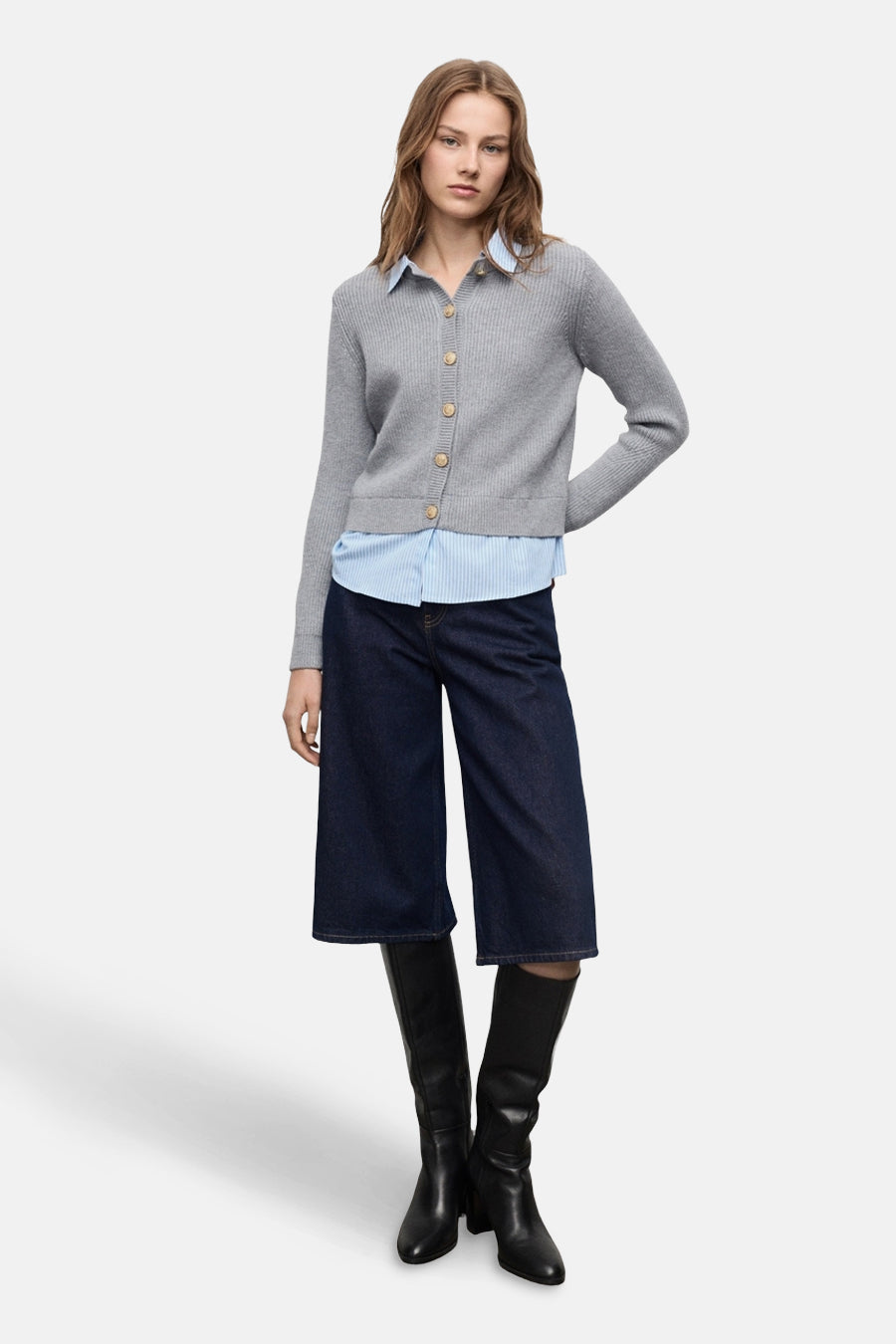Cardigan gris de MANGO, porté sur une chemise rayée bleu ciel, avec un pantalon large en denim et des bottes noires.