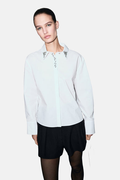 Chemise blanche à manches longues de MANGO, avec col orné de pierres décoratives, portée avec un short noir et des collants noirs.
