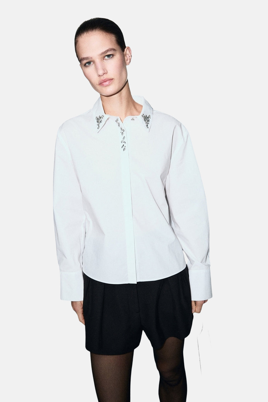 Chemise blanche à manches longues de MANGO, avec col orné de pierres décoratives, portée avec un short noir et des collants noirs.
