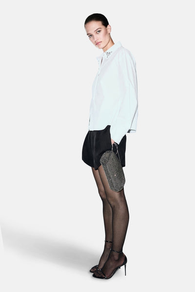 Chemise blanche à manches longues de MANGO, ornée de détails décoratifs au niveau du col, portée avec un short noir et des collants noirs.
