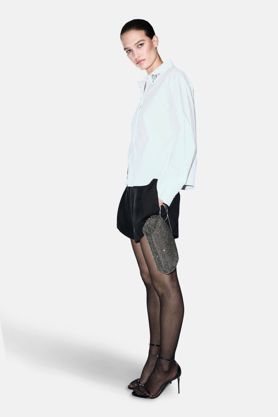 Chemise blanche à manches longues de MANGO, ornée de détails décoratifs au niveau du col, portée avec un short noir et des collants noirs.
