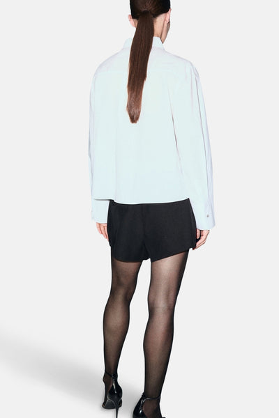 Chemise blanche à manches longues de MANGO, vue de dos, portée avec un short noir et des collants.