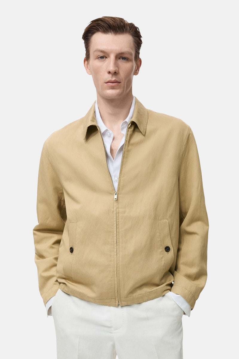 Korte beige jas van MANGO, met ritssluiting en zakken met knopen, gedragen over een wit overhemd en een witte broek.