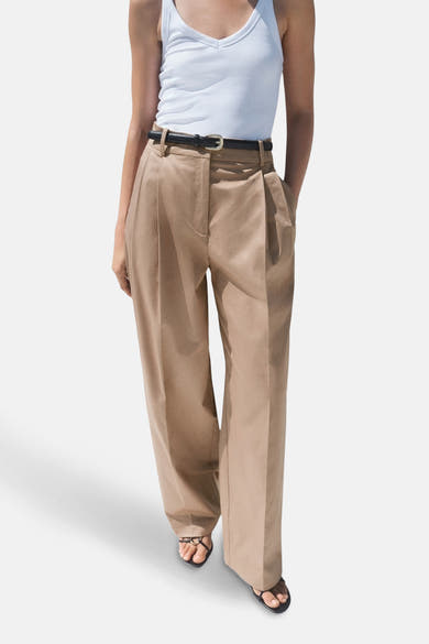Pantalon beige de MANGO, large avec plis, porté avec une ceinture noire et des sandales.
