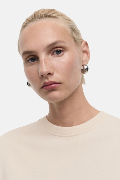 T-shirt beige à manches courtes de MANGO, porté avec des boucles d'oreilles rondes argentées.