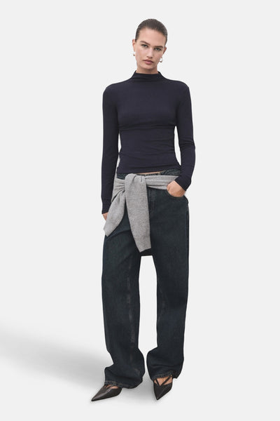 T-shirt bleu à manches longues de MANGO, porté avec un pull gris noué à la taille et un jean ample.

