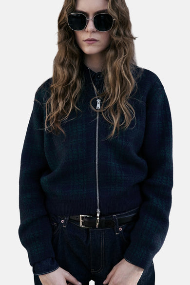 Gilet multicolore de MANGO, à fermeture éclair argentée, porté avec une ceinture en cuir et des lunettes de soleil.
