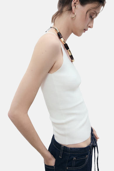 Witte tanktop van MANGO, gedragen met een ketting met grote gouden en bruine kralen en een low-rise spijkerbroek.