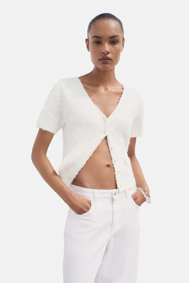 Cardigan blanc en maille texturée par MANGO, porté ouvert avec un jean blanc et un bracelet transparent.