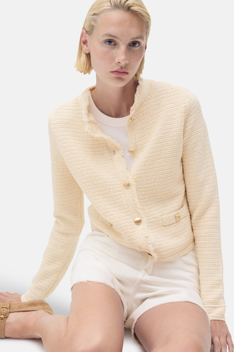 Ecru gebreid vest met textuur van MANGO, met rafelrand en gouden knopen, gedragen over een wit T-shirt en een witte korte broek.