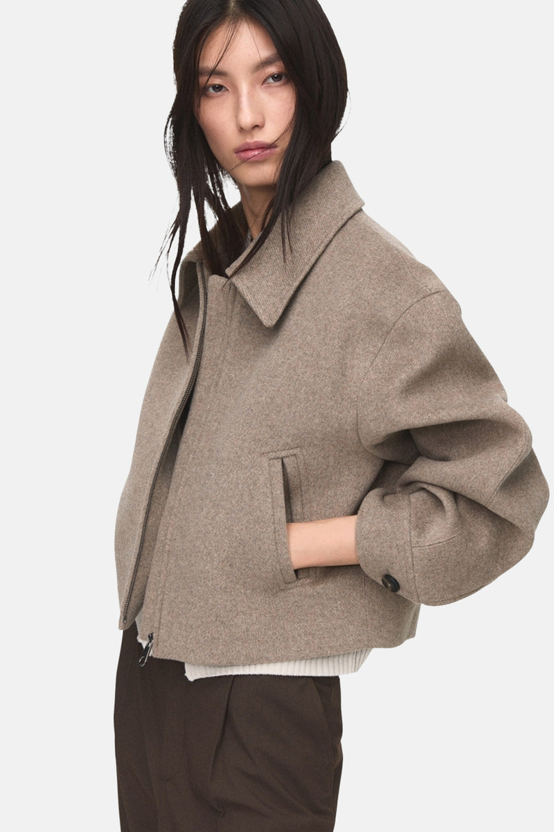Korte beige jas van MANGO, met ritssluiting en zakken, gedragen met een bruine broek.