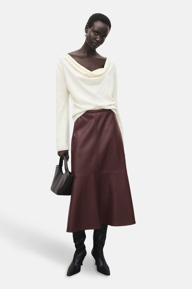 Jupe longue bordeaux en similicuir de MANGO, portée avec un pull blanc et des bottes noires.