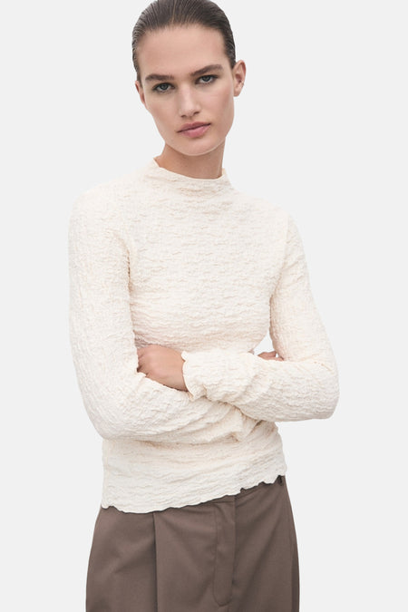 T-shirt à manches longues beige de MANGO, en tissu texturé, porté avec un pantalon marron.
