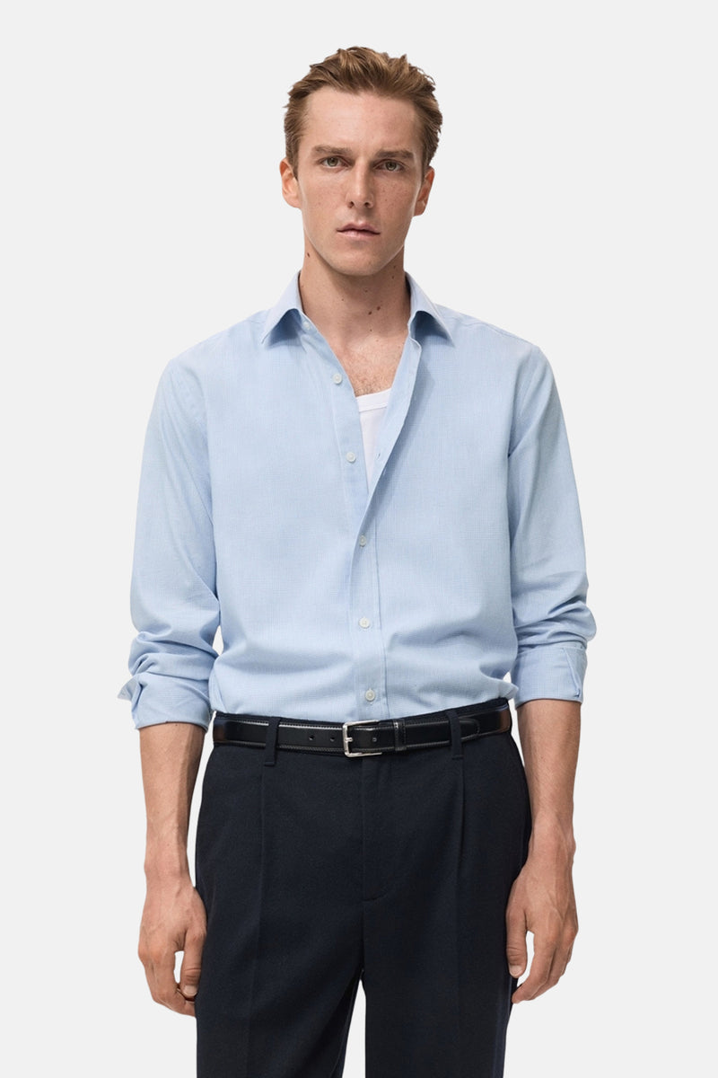 Chemise à manches longues bleue de MANGO, portée avec un pantalon de costume noir et une ceinture en cuir.
