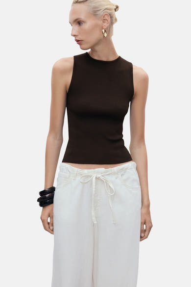 Top sans manches brun de MANGO, porté avec un pantalon blanc à cordon et des bracelets noirs.
