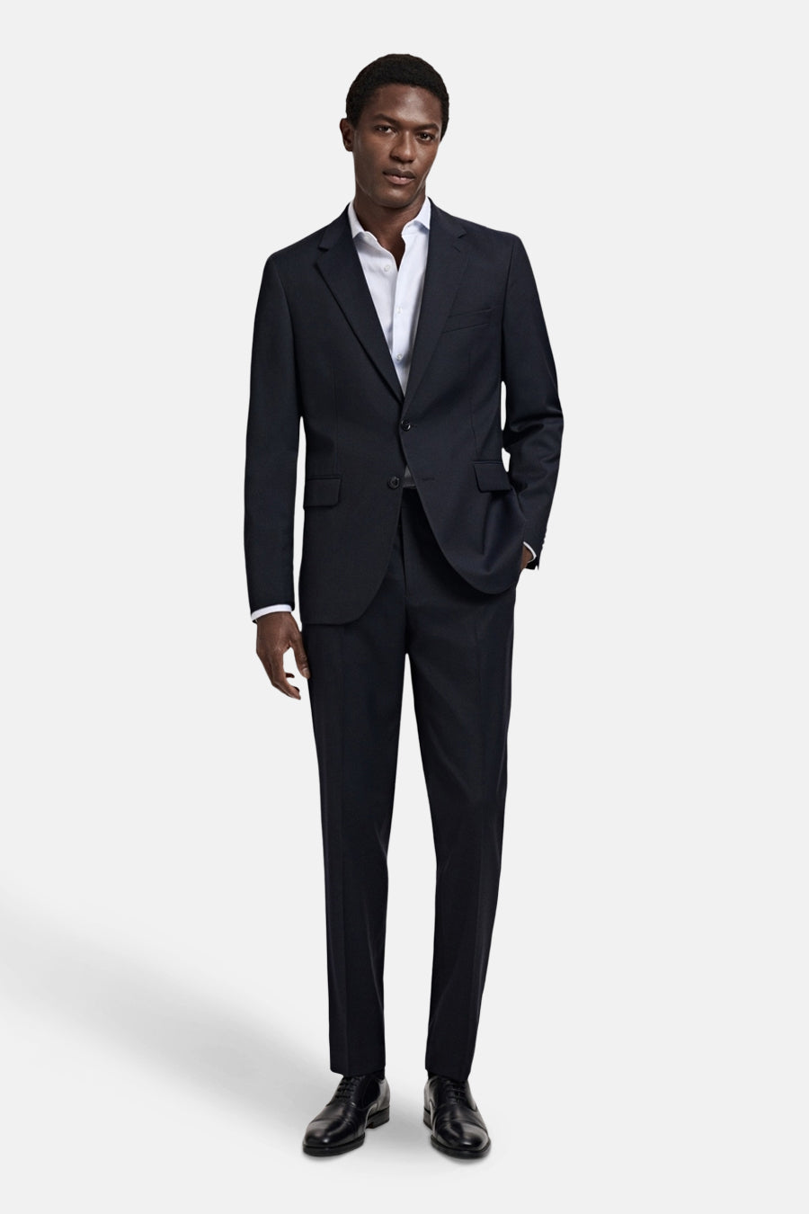 Blauwe blazer van MANGO, slim fit, twee knopen, ingesneden revers en klepzakken. Gedragen met een bijpassende broek, wit overhemd en zwarte schoenen.
