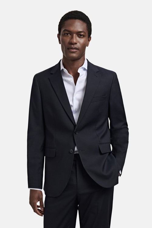 Blazer bleu de MANGO porté par un homme avec une chemise blanche rayée ouverte et la main dans la poche.
