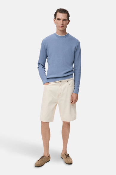 Pull à col rond bleu de MANGO, porté avec un short beige et des chaussures marron.