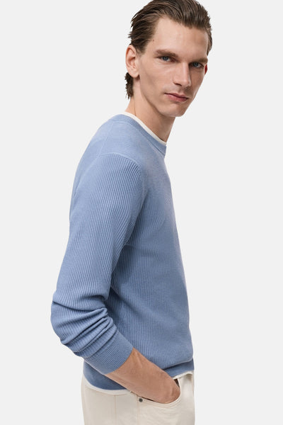 MANGO blauwe rondehals trui in ribgebreide stof, gedragen met een gebroken wit T-shirt en een beige broek, zijwaarts bekeken.
