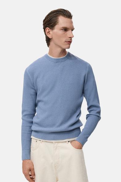 Pull bleu à col rond de MANGO, avec une texture côtelée, porté sur un t-shirt blanc et un pantalon clair.