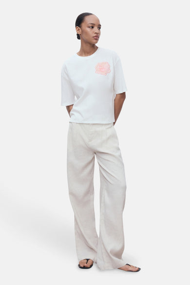 T-shirt blanc à manches courtes de MANGO, avec un motif floral rose sur le devant, porté avec un pantalon ample clair.
