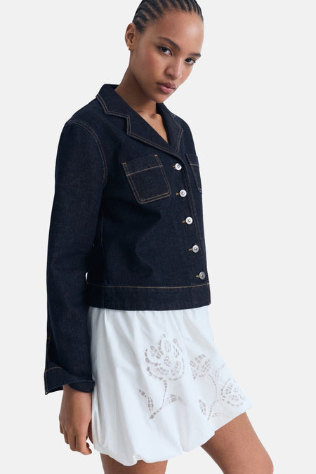 Veste en jean bleu foncé de MANGO, avec surpiqûres contrastées et boutons blancs, portée sur une robe blanche brodée.
