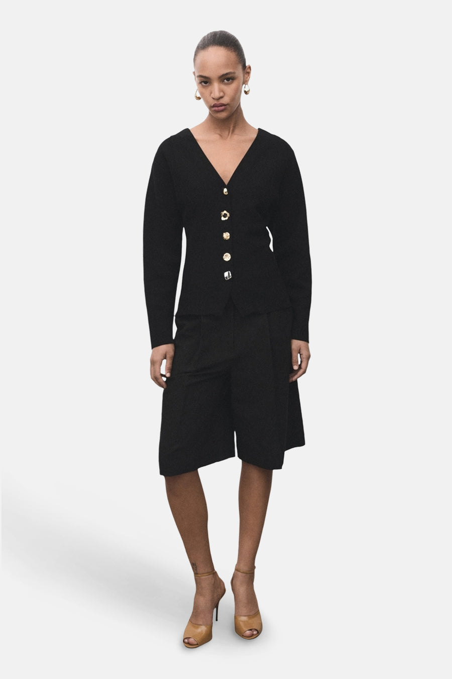 Cardigan noir par MANGO, avec boutons dorés, porté avec un short noir ample et des talons hauts beiges.