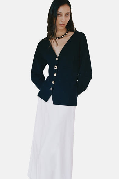 Cardigan noir by MANGO, à encolure en V, avec des boutons dorés, porté avec une jupe blanche.