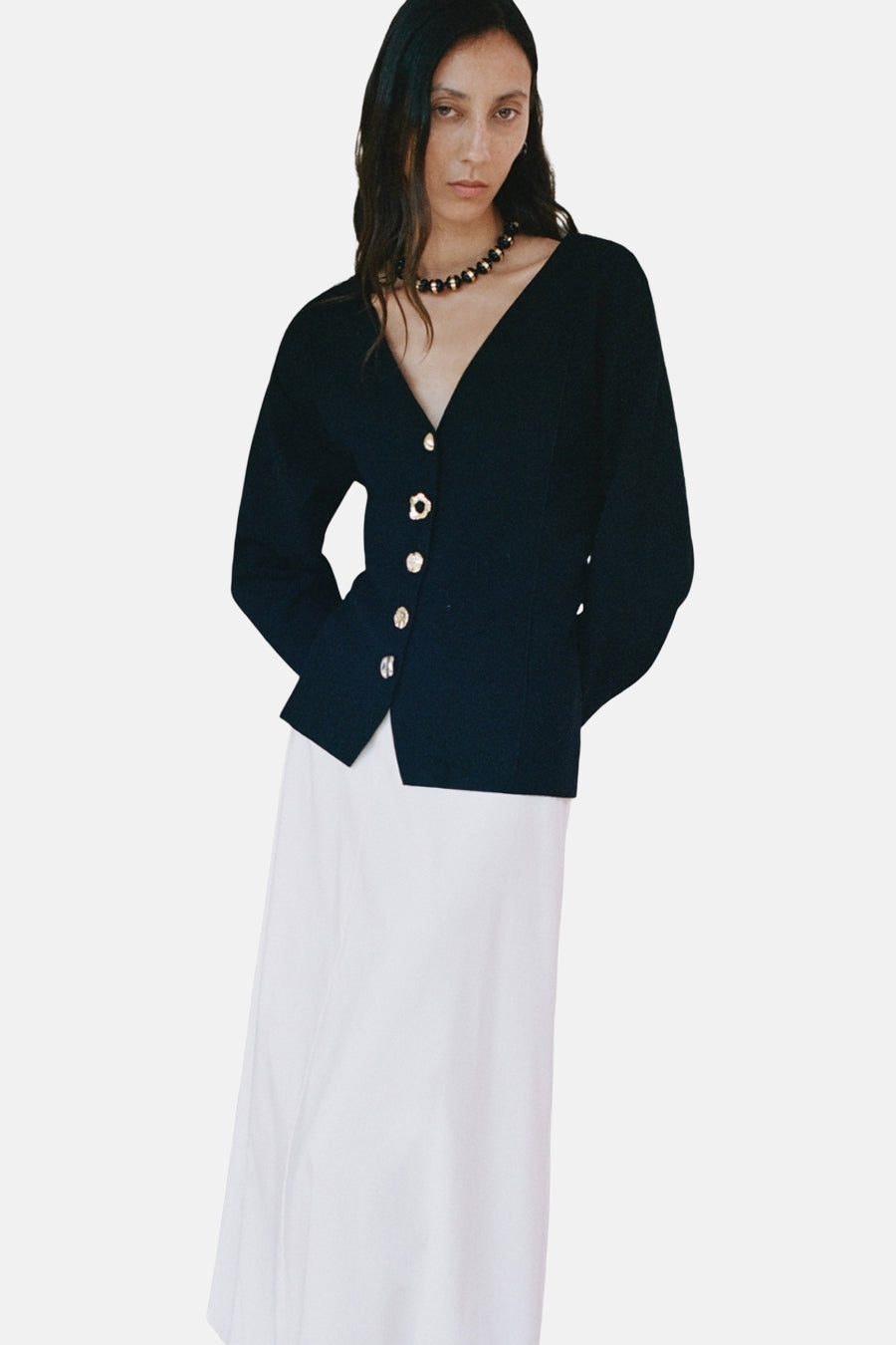 Cardigan noir by MANGO, à encolure en V, avec des boutons dorés, porté avec une jupe blanche.