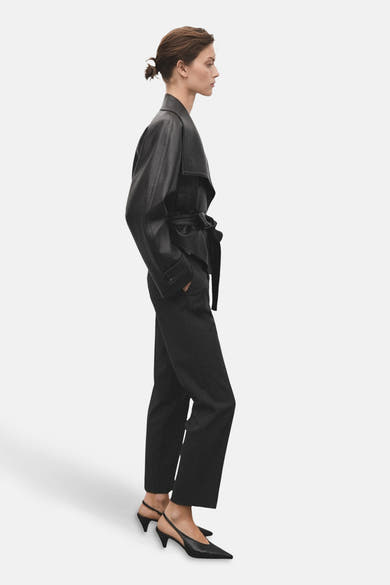 Pantalon gris de MANGO, porté avec une veste en cuir ceinturée et des escarpins noirs à bride.