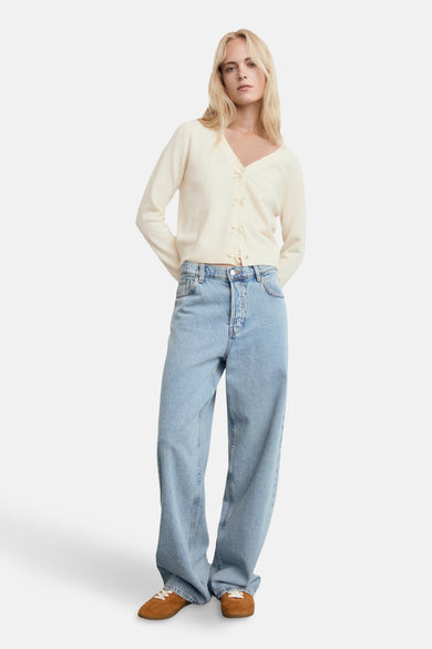 Cardigan écru de MANGO, coupe décontractée avec petits nœuds décoratifs, porté avec un jean ample et des chaussures marron.
