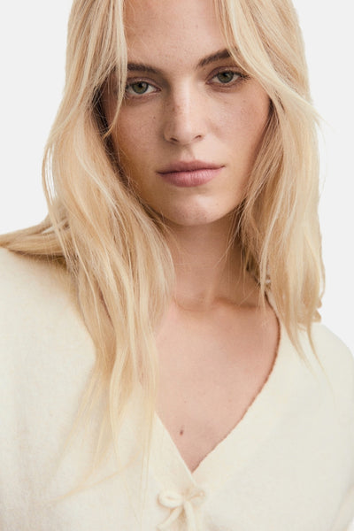 Ecru vest van MANGO, met een zachte textuur en een decoratieve strik, gedragen met blond haar en een lichte huidskleur.