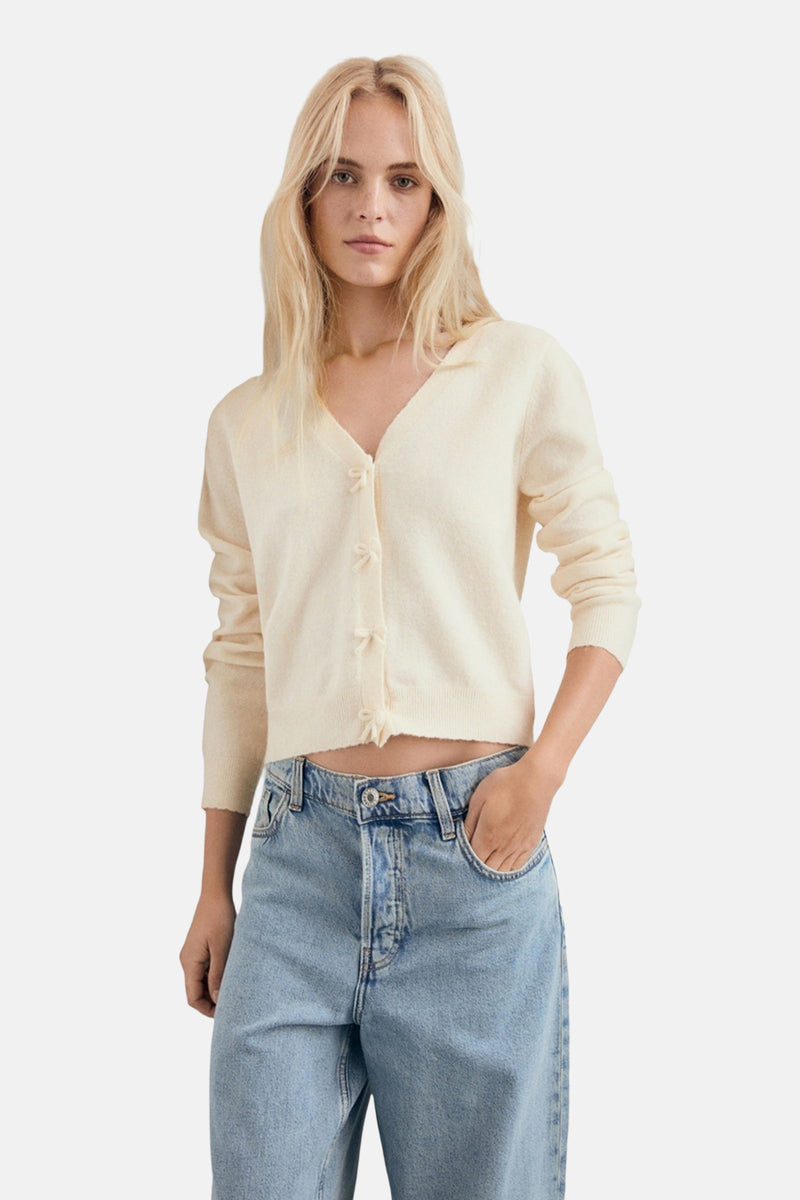 Cardigan écru de MANGO avec des petits nœuds décoratifs, porté avec un jean taille haute.
