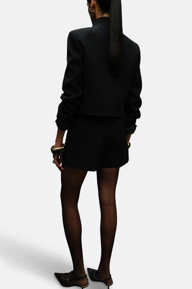 Blazer noir de MANGO, porté avec un short noir, des collants noirs et des escarpins à talons aiguilles.