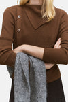 Pull à col rond brun de MANGO avec boutonnage asymétrique, porté avec une écharpe grise.
