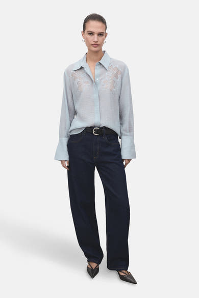 Chemise bleue à manches longues de MANGO, ornée de broderies florales sur le devant, portée avec un jean et une ceinture noirs.
