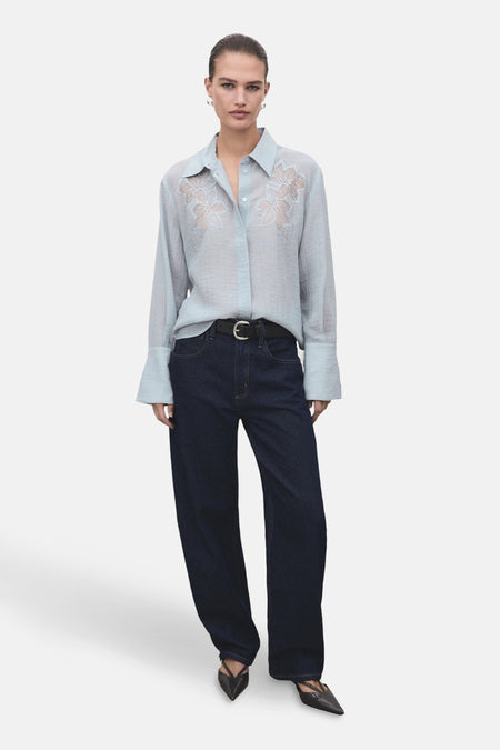 Chemise bleue à manches longues de MANGO, ornée de broderies florales sur le devant, portée avec un jean et une ceinture noirs.
